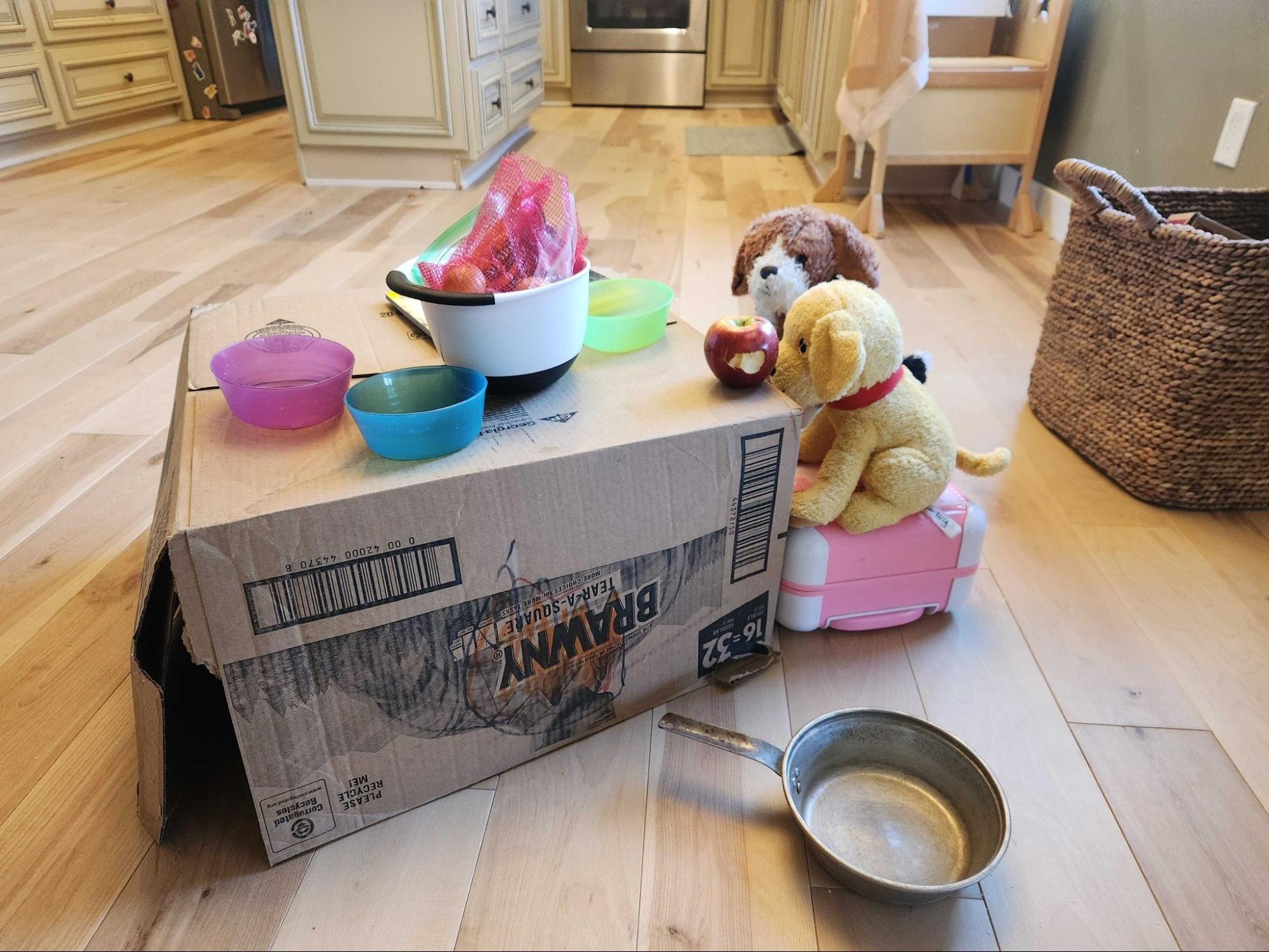Photo of two stuffed animals sitting at a table made out of a recycled Brawny box on a wood kitchen floor. The box has an apple on it as well as some dishes and vegetables in a bag. There is a frying pan on the floor in front of the box.