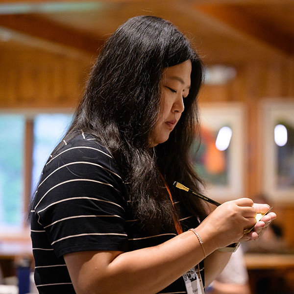 A person with long black hair, wearing a black and white striped shirt, stands indoors, focusing intently while writing with a pen. The background shows a wooden cabin-like interior, with soft lighting and blurred artwork on the walls.