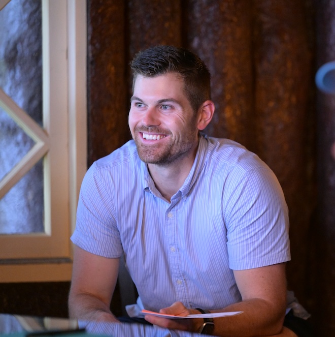 A man with short brown hair and a beard, wearing a light blue button-up shirt, smiles while sitting indoors. The background features a rustic wooden wall and soft lighting, with part of a window visible on the left.