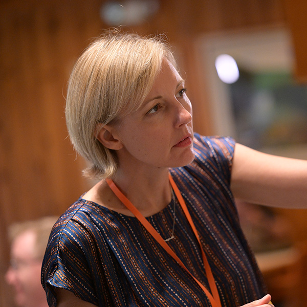 A woman with short, blonde hair and a focused expression looks off to the side. She wears a blue and orange patterned top and an orange lanyard, suggesting a professional or conference setting.