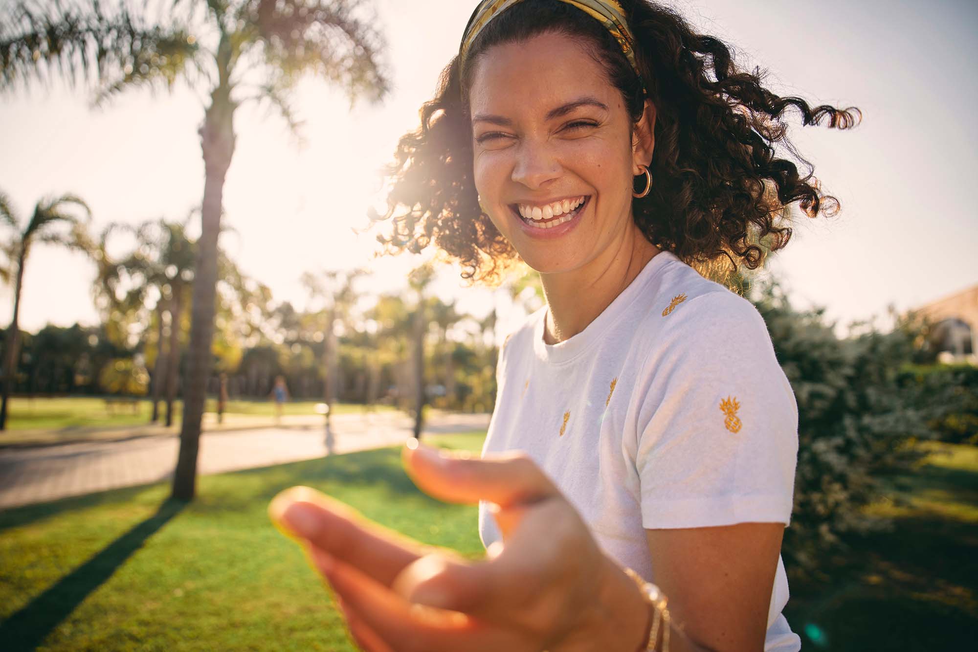 A woman with curly hair, wearing a white shirt with small yellow pineapple prints, is smiling and reaching toward the camera. She is outdoors in a park with palm trees and greenery, and the sunlight creates a warm, bright atmosphere. She is also wearing a headband and hoop earrings.