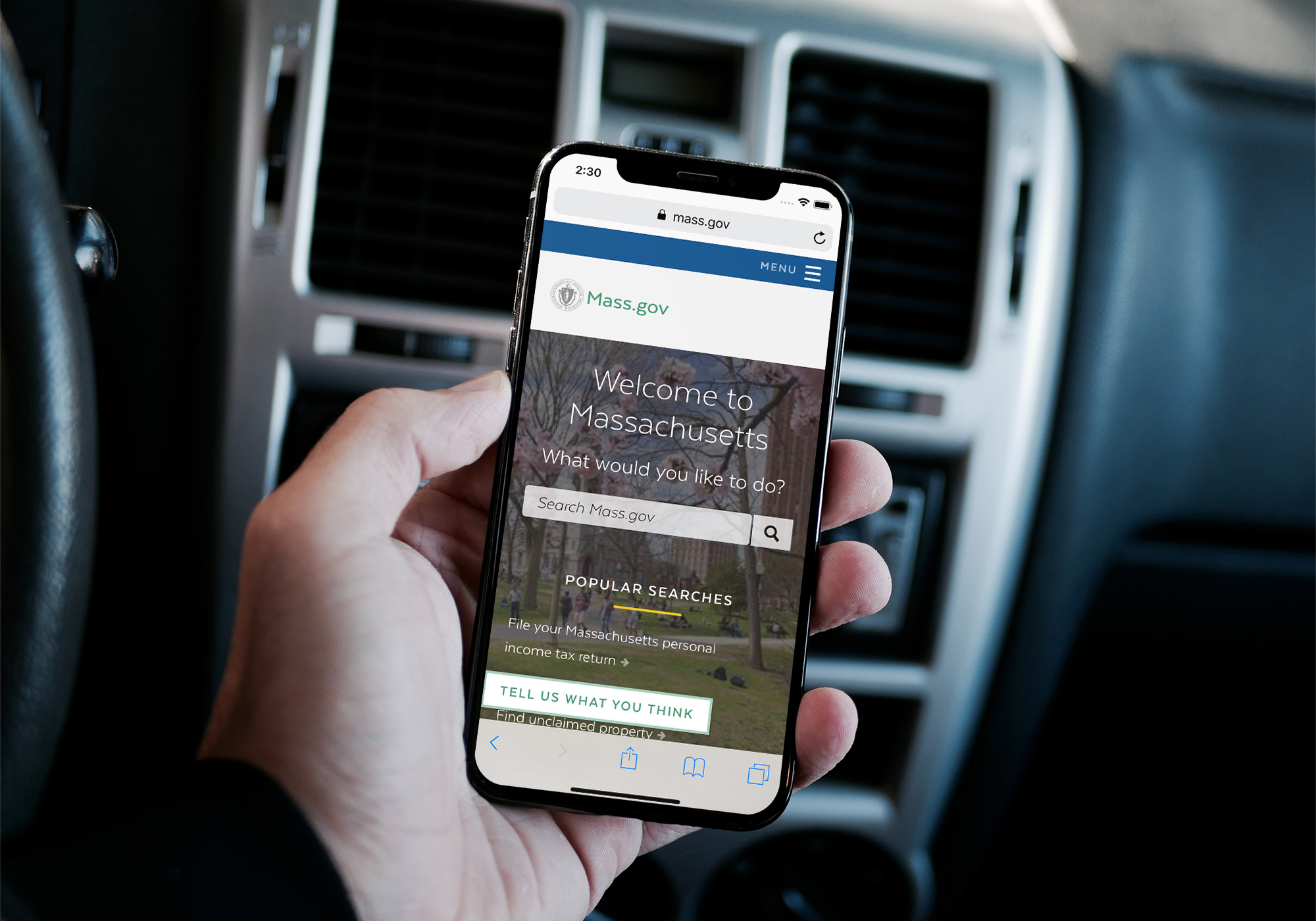 Hand holding a smartphone showing the Mass.gov website with a “Welcome to Massachusetts” message and search bar, inside a car.