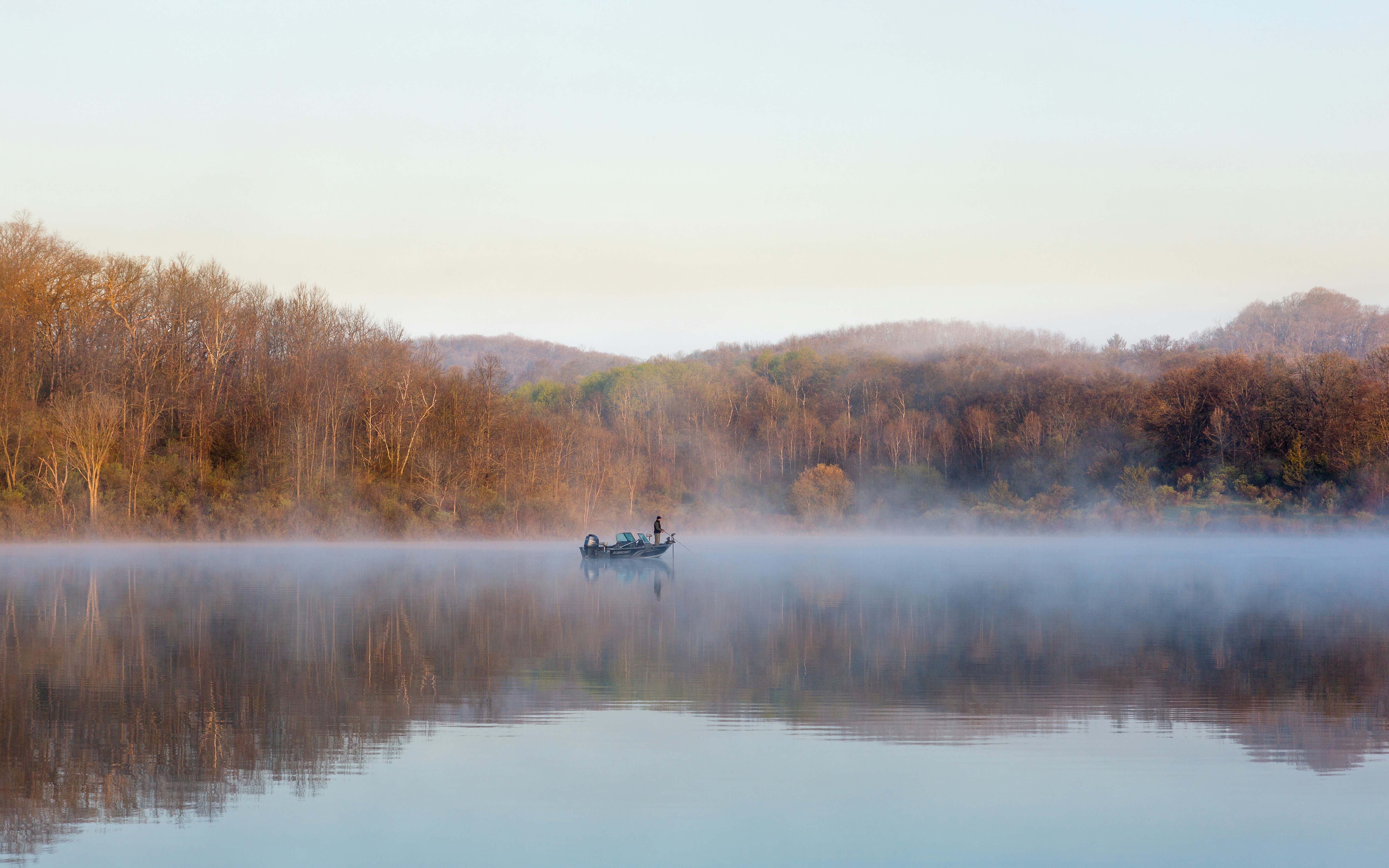 A lone fisherman in a boat on a misty lake, surrounded by bare trees and calm reflections in the early morning light.