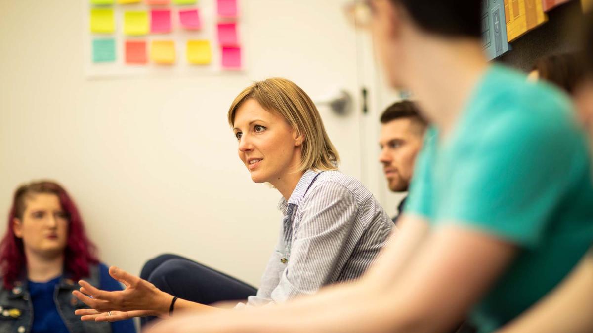 Woman sitting in a group with other people holding out her hands. There is a white wall with stickies on it in the background.