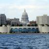 A scenic view of the Madison, Wisconsin skyline, featuring the iconic Wisconsin State Capitol building rising majestically in the center. In the foreground, the Monona Terrace Community and Convention Center stretches along the shore of Lake Monona, its curved facade reflecting the blue sky and clouds.